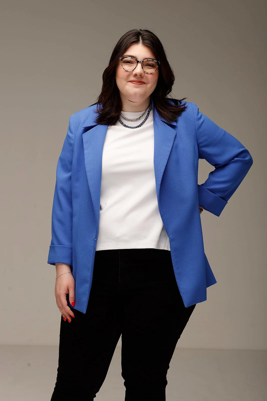 A woman with shoulder-length dark hair, glasses, and a slight smile, wearing a blue blazer over a white shirt, black pants, and layered necklaces, standing with her right hand on her hip against a plain beige background.