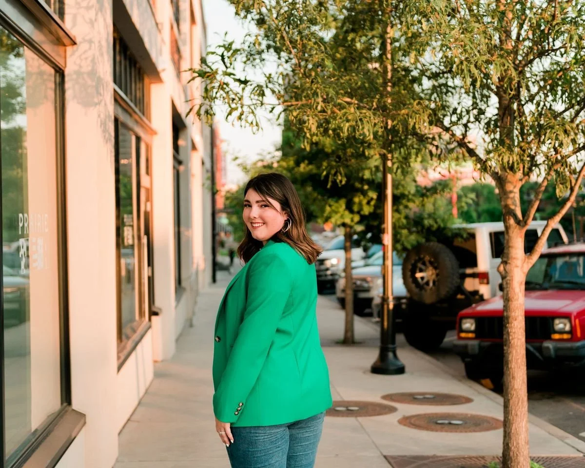 Young woman with dark hair smiling and turning to face the camera, wearing a bright green blazer and blue jeans, standing on a sidewalk next to a building with large windows, with trees and parked cars in the background, during sunset.