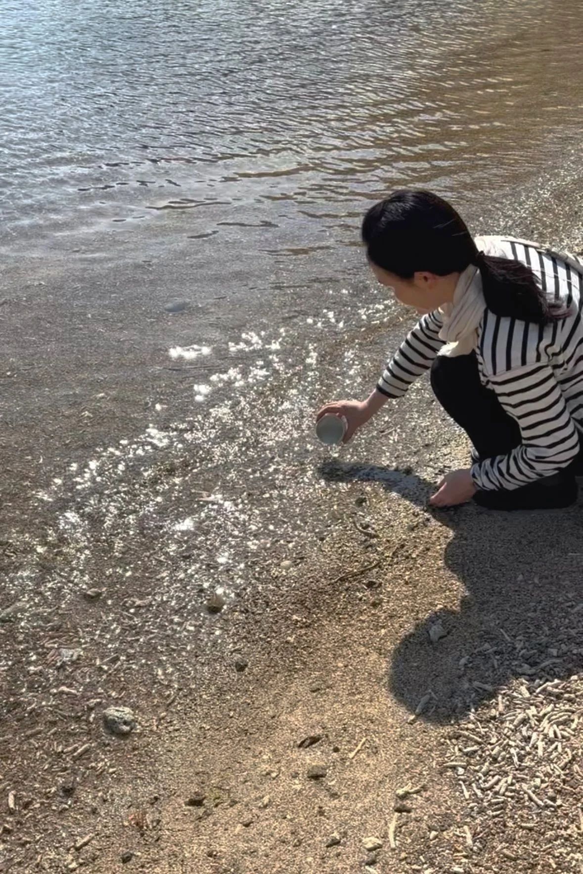Woman crouching on sandy beach, pouring water from a cup into the shallow surf