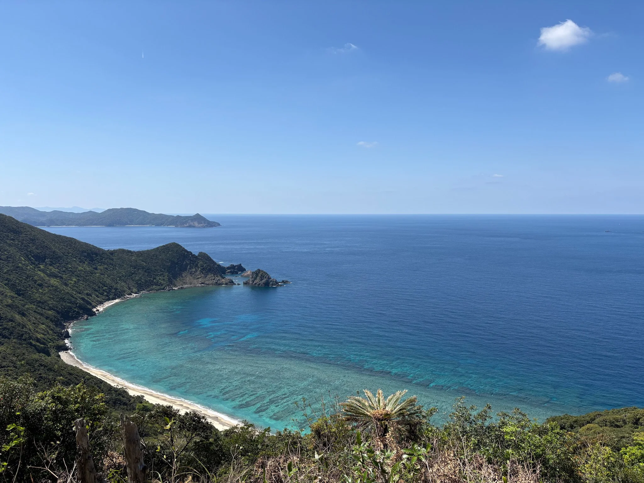 A scenic view of a crescent-shaped beach with clear blue water, lush green hills surrounding it, and a mostly clear sky with a few small clouds.