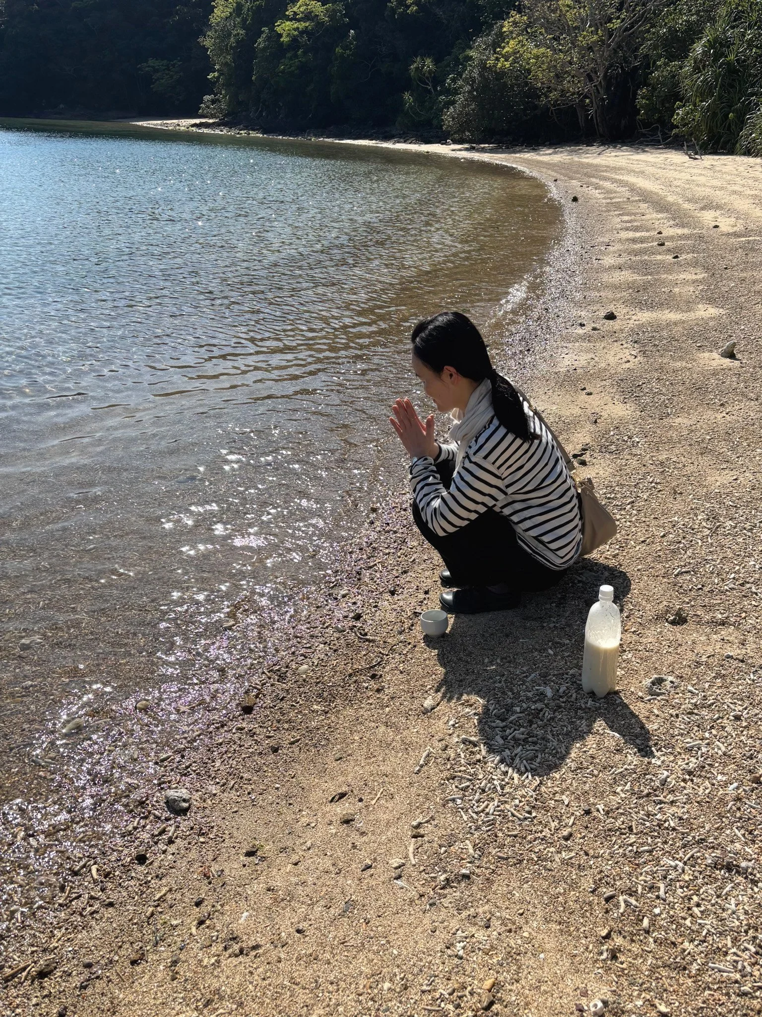 A woman crouches on a sandy beach near the water, with her hands together as if praying or being reflective, beside a bottle of milk and small bowls.