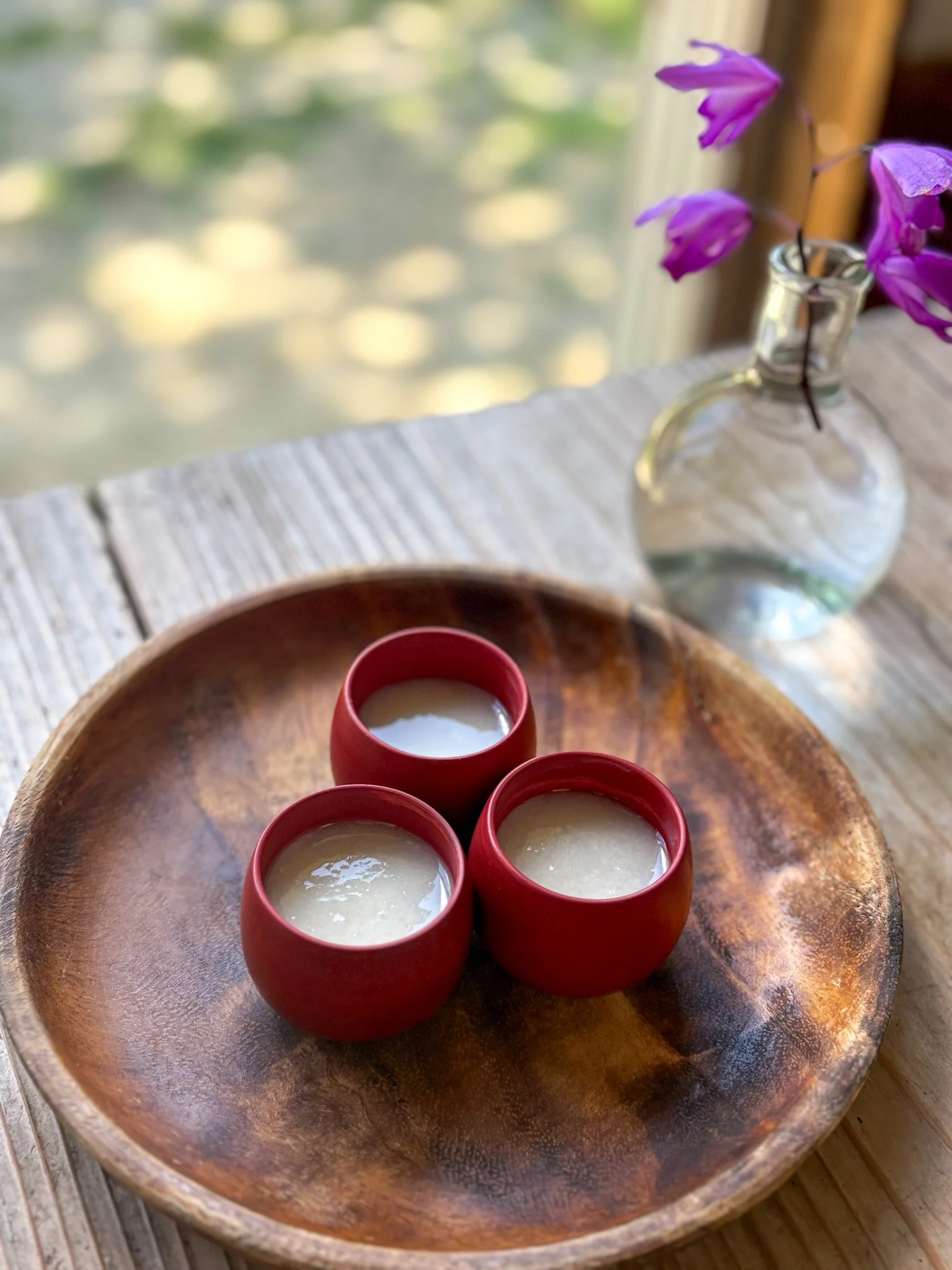 Three small red bowls filled with a white substance on a wooden plate beside a glass vase with purple flowers, on a wooden table near a window, blurred green outdoor background.