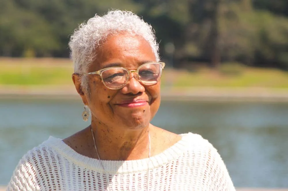 Smiling elderly Black woman wearing glasses and a white sweater outdoors by a lake