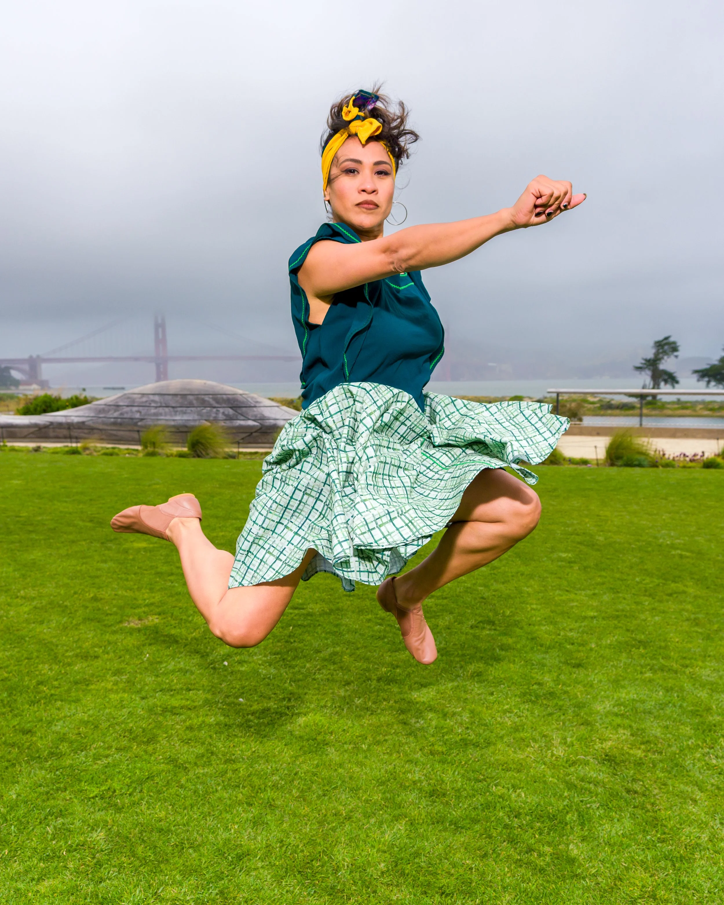 Dancer in a yellow headband, teal top, and checkered skirt leaping across a grassy field near a waterfront bridge
