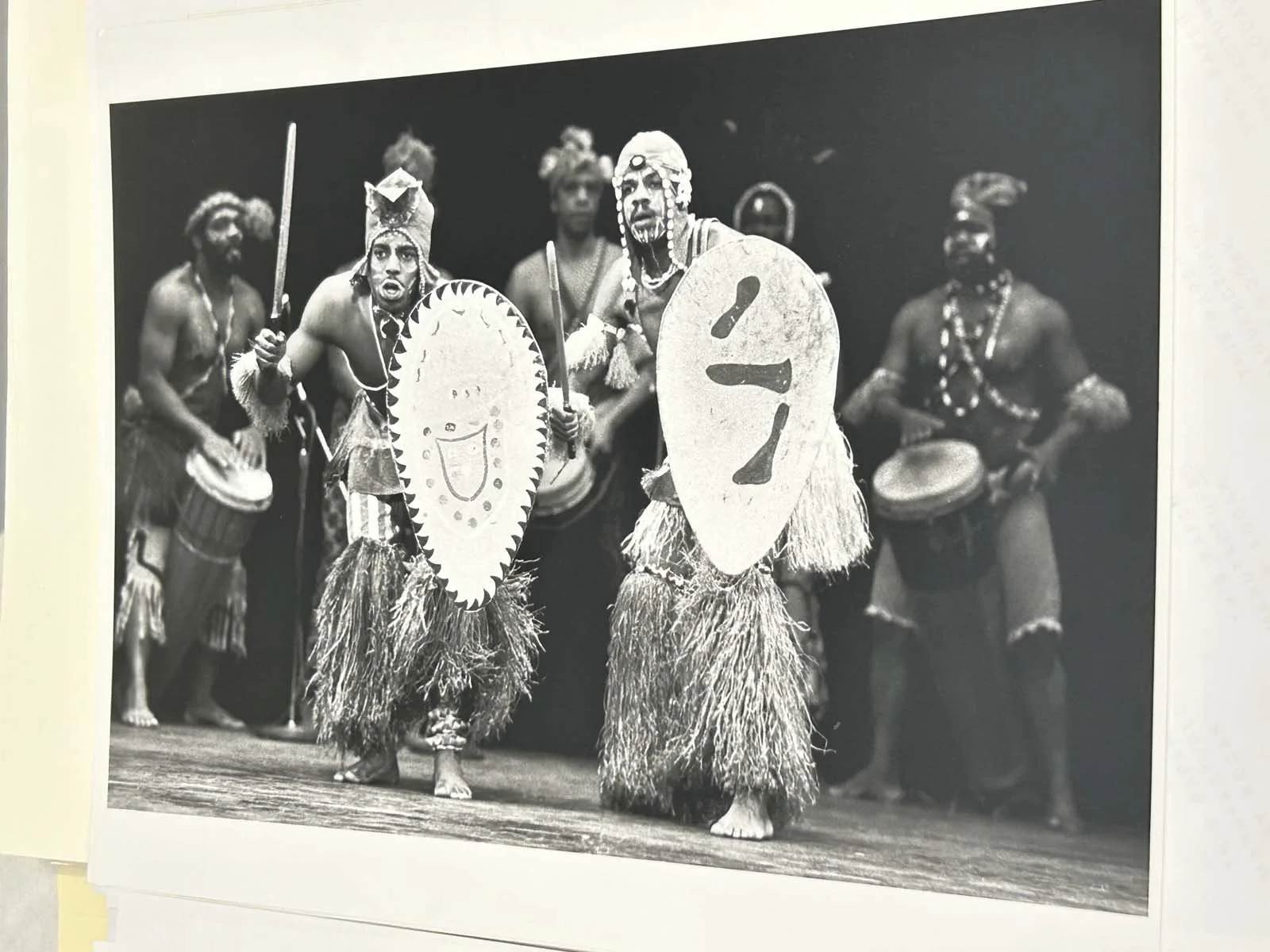 Black-and-white theatrical performance featuring two costumed shield-bearing warriors, drummers, and performers on stage