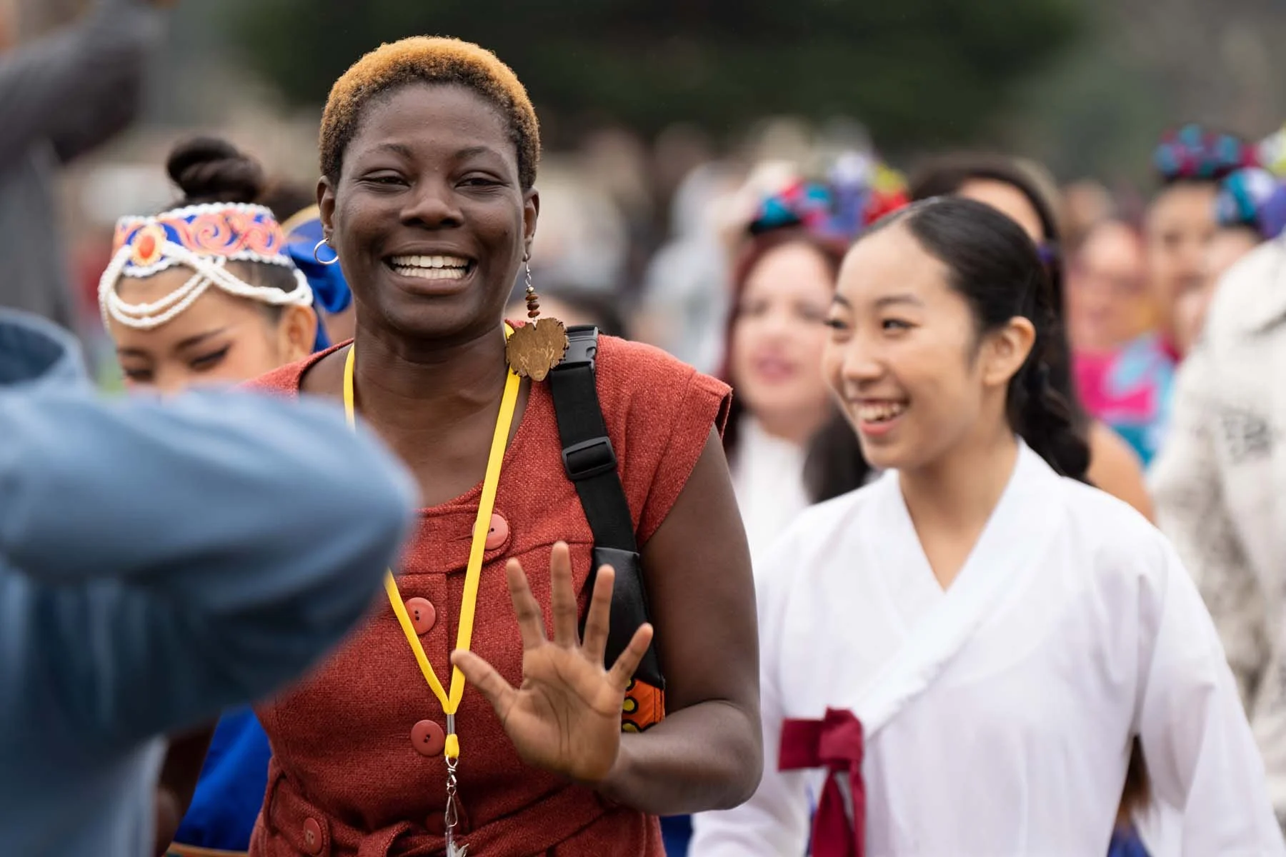 Smiling woman waving among a diverse outdoor festival crowd, with a woman in a white kimono beside her