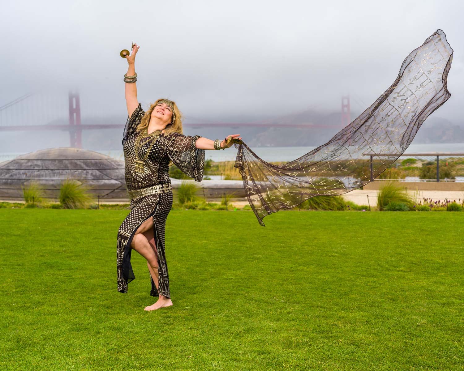 Woman in patterned black outfit dancing with a flowing scarf on a lawn near San Francisco’s Golden Gate Bridge