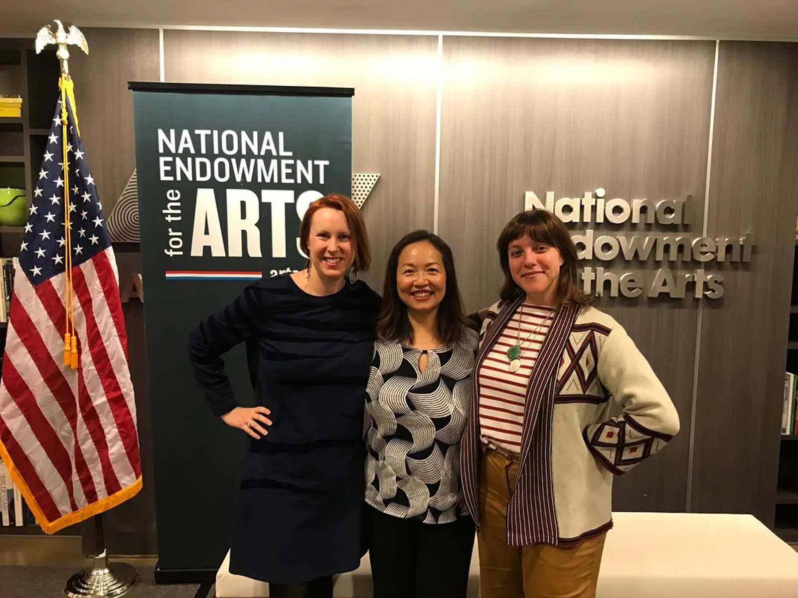 Three women pose beside a U.S. flag and National Endowment for the Arts sign.