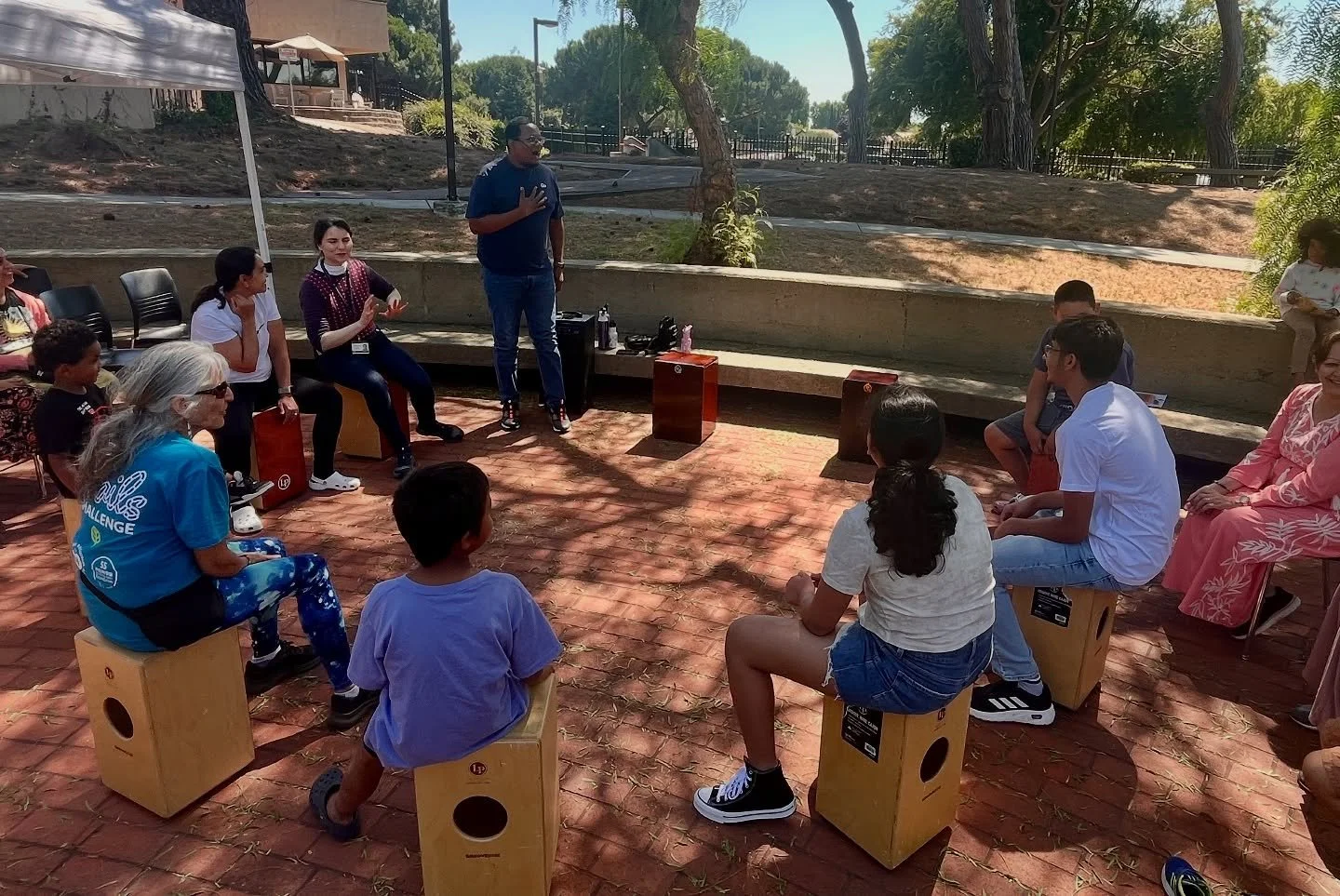Outdoor group drumming circle with participants seated on cajón percussion boxes in a shaded park
