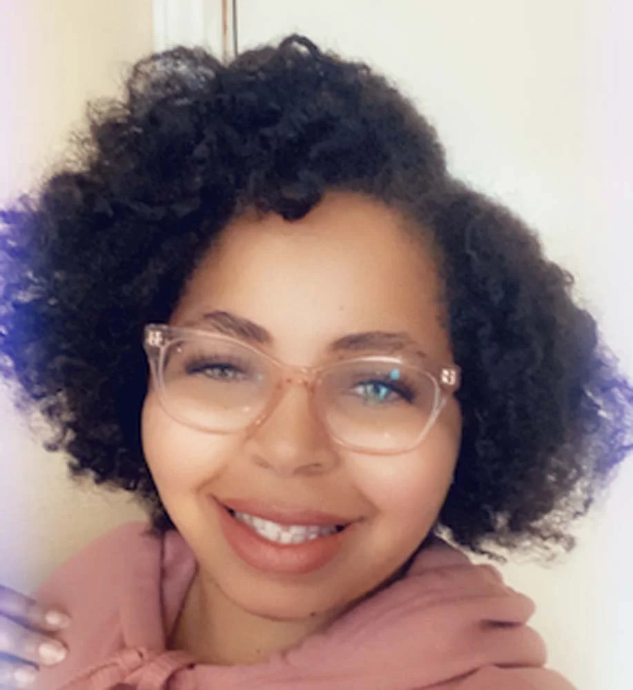 Smiling woman with curly hair wearing clear-framed glasses and a pink hoodie