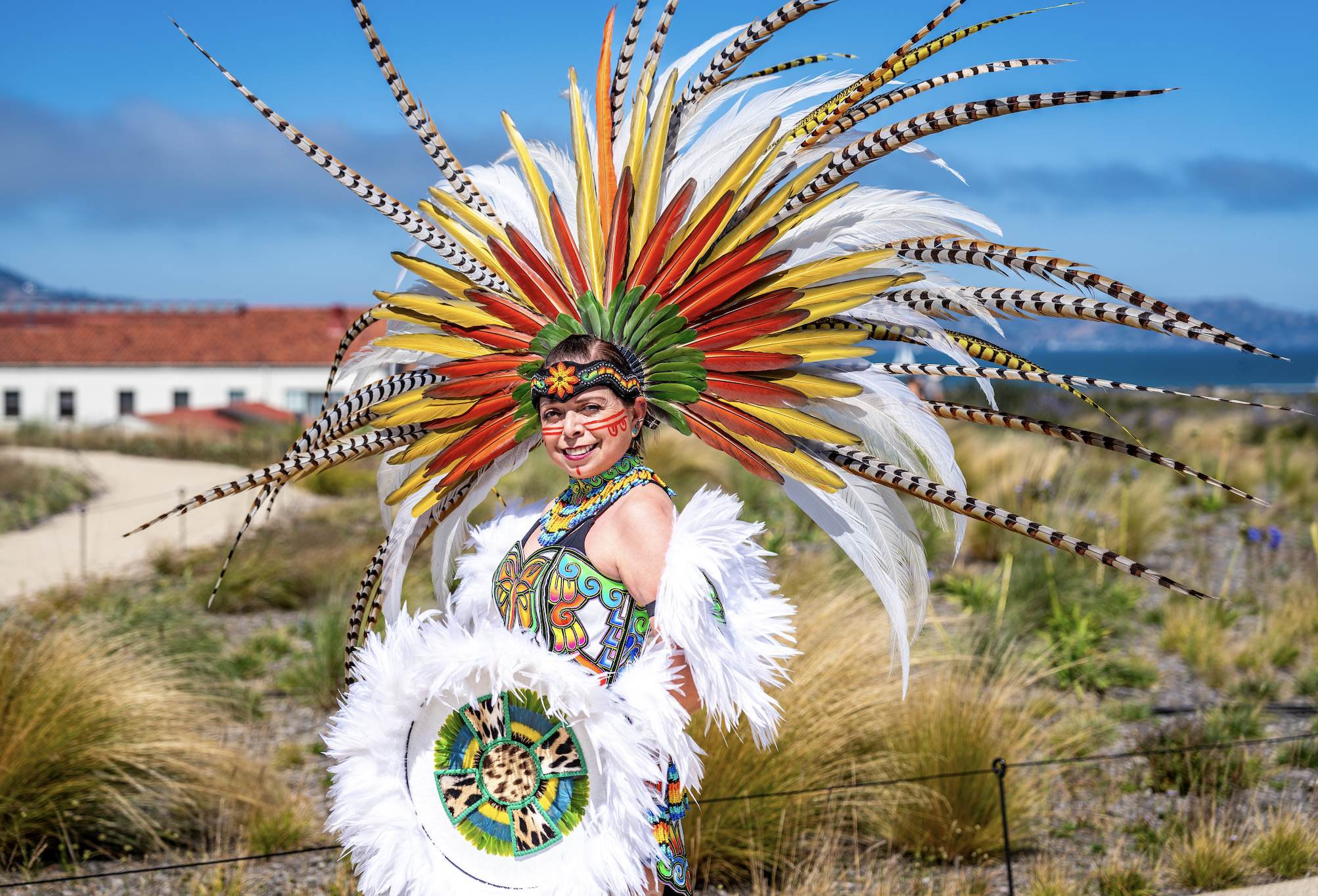 Woman in colorful feathered headdress and traditional costume posing outdoors by the coast