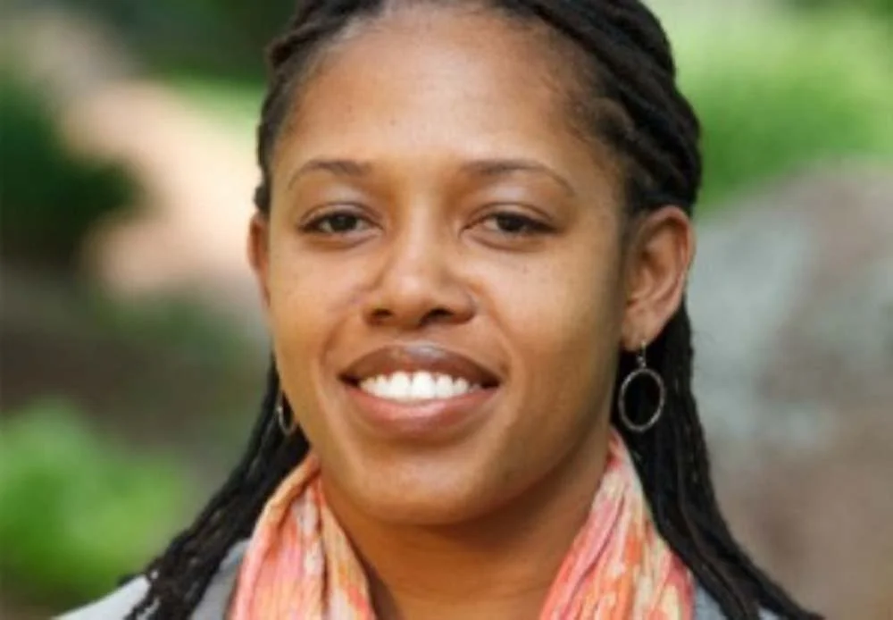 Portrait of a smiling Black woman with braided hair, hoop earrings, and a pink scarf outdoors