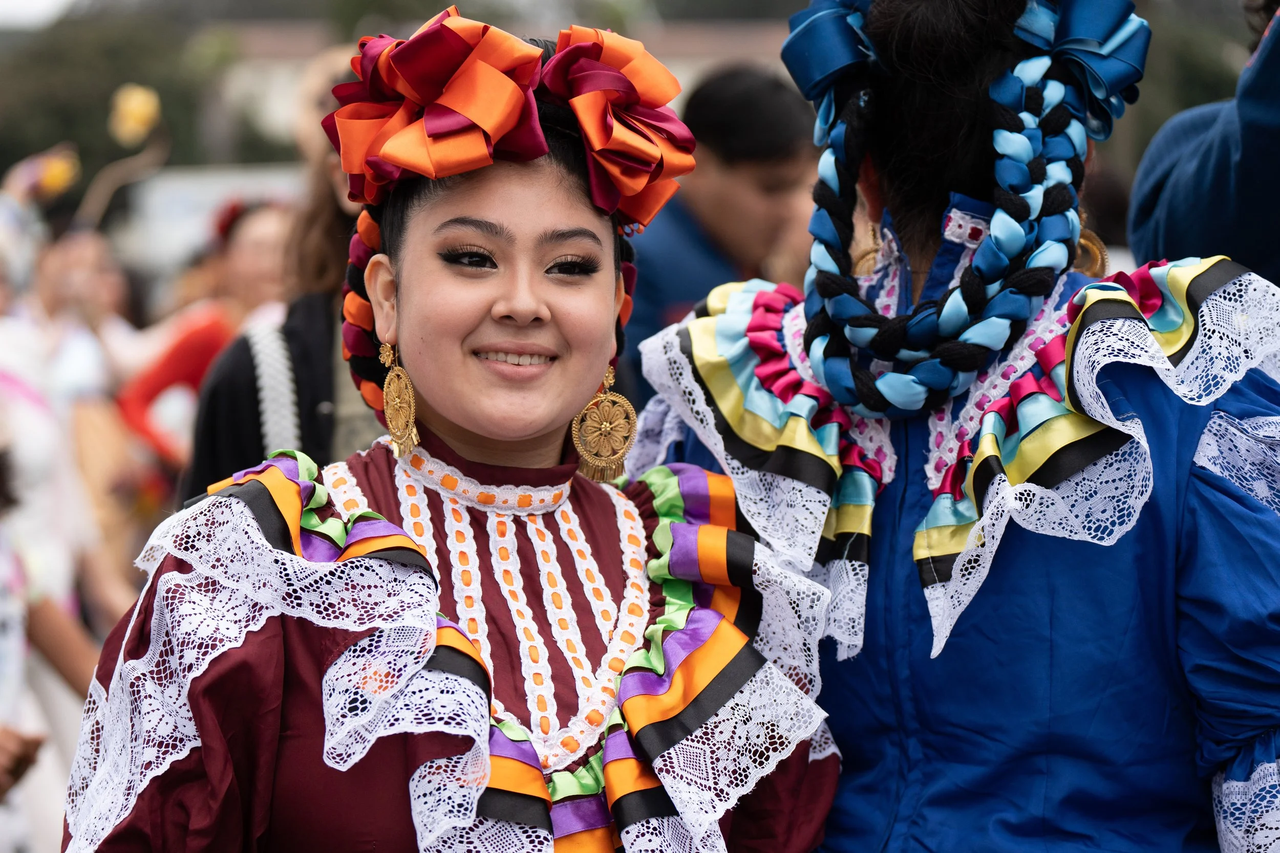 Smiling dancers in colorful traditional folkloric dresses with lace, ribbons, and decorative hair bows at a cultural festival