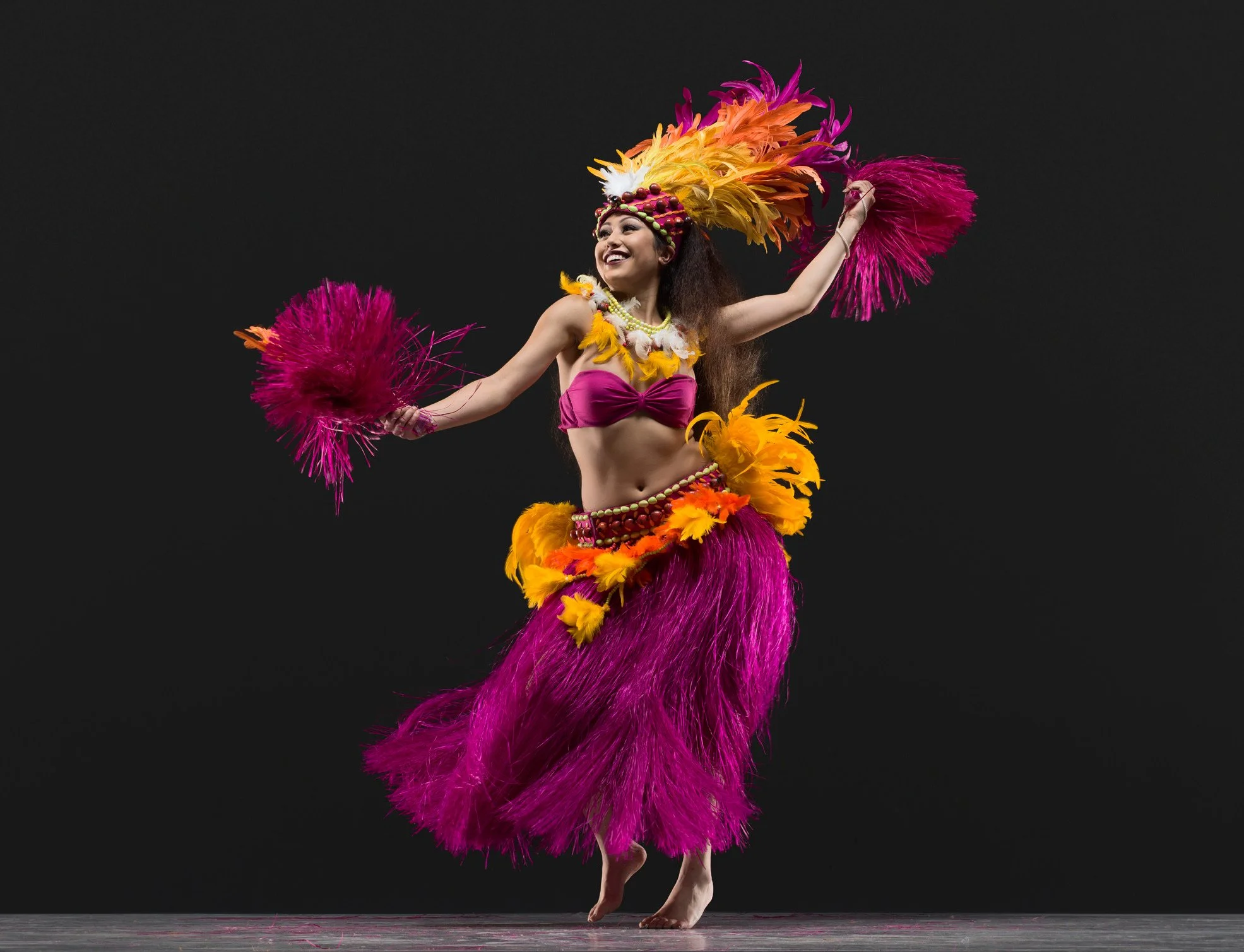 Smiling Polynesian dancer in a vibrant feathered costume performing with colorful pom-poms against a black background