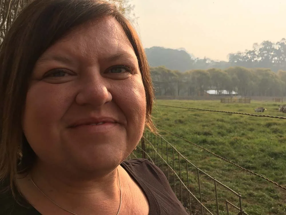 Woman taking a selfie outdoors beside a fenced grassy field at sunrise