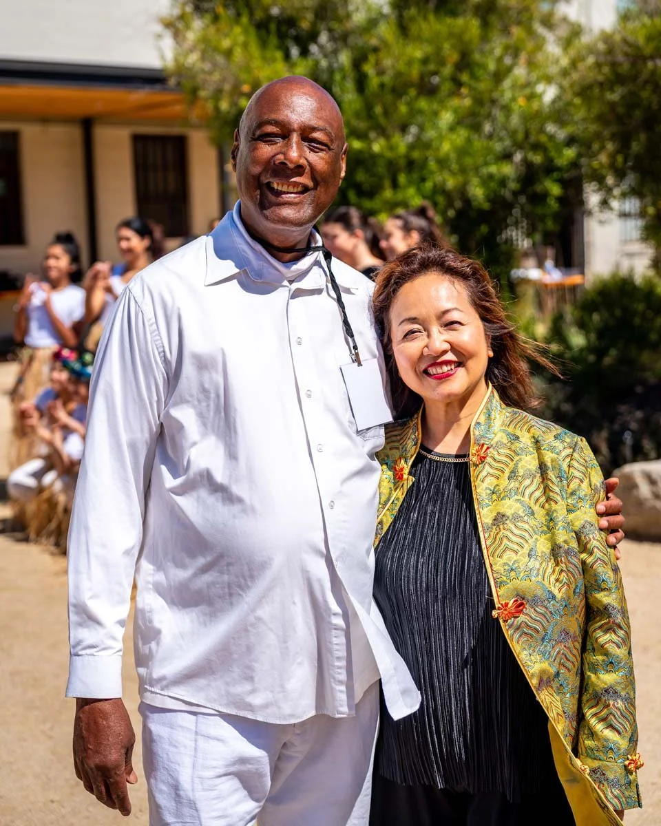 Smiling couple posing outdoors at a sunny gathering, with people and trees in the background