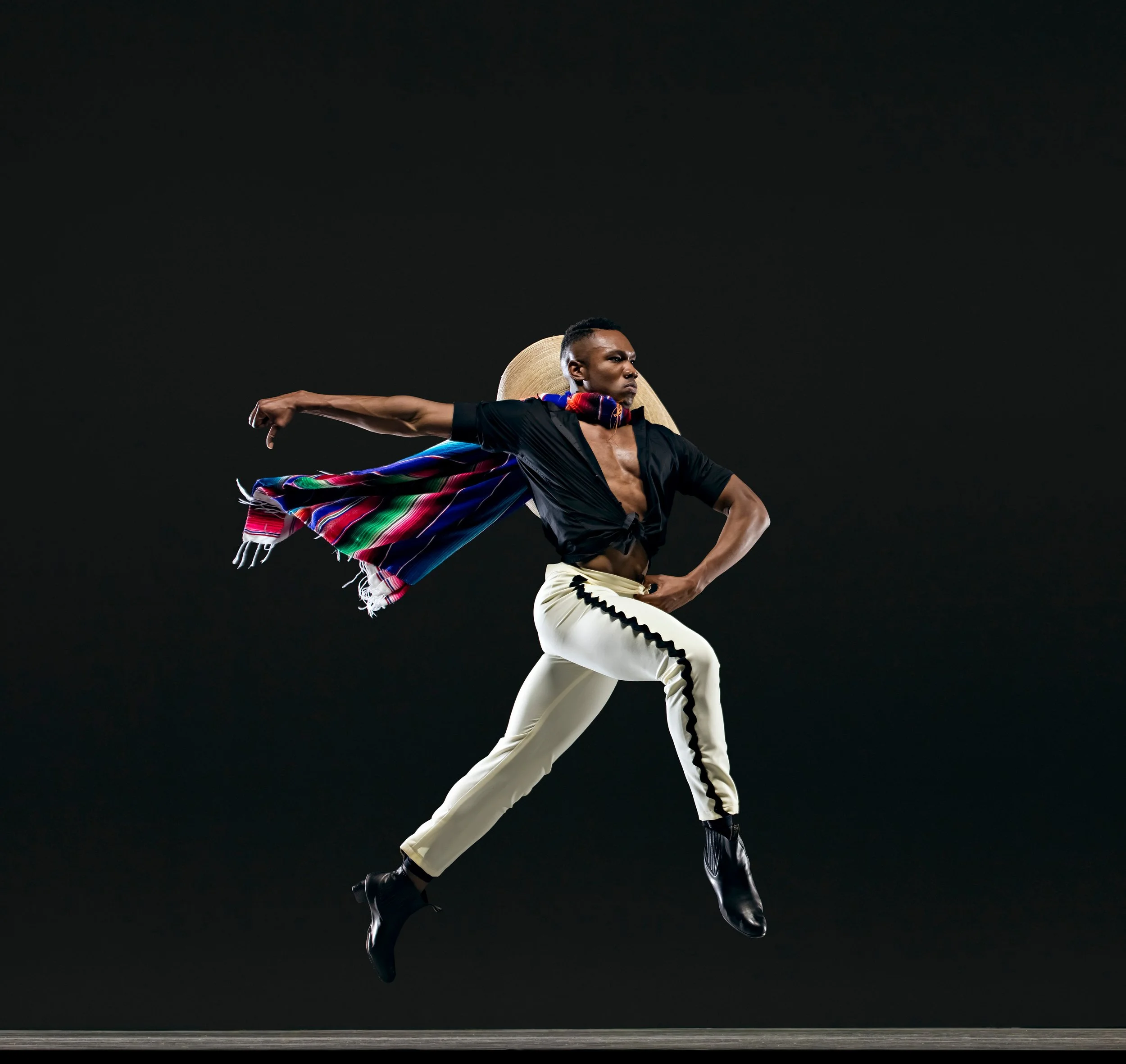 Dancer leaping in black and white clothing with a colorful scarf against a black background