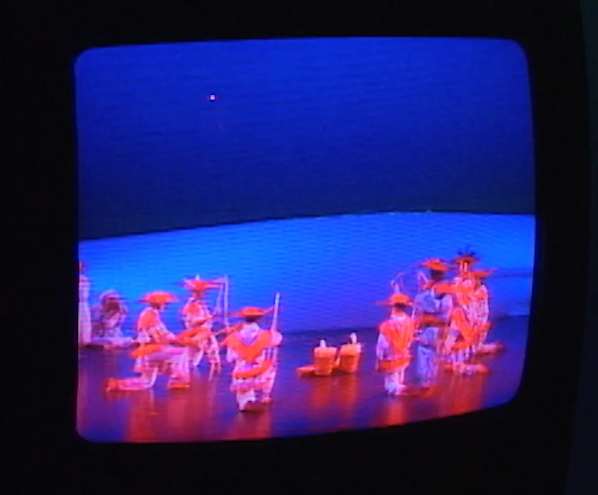 Colorful stage performance featuring dancers in orange costumes and hats under blue lighting