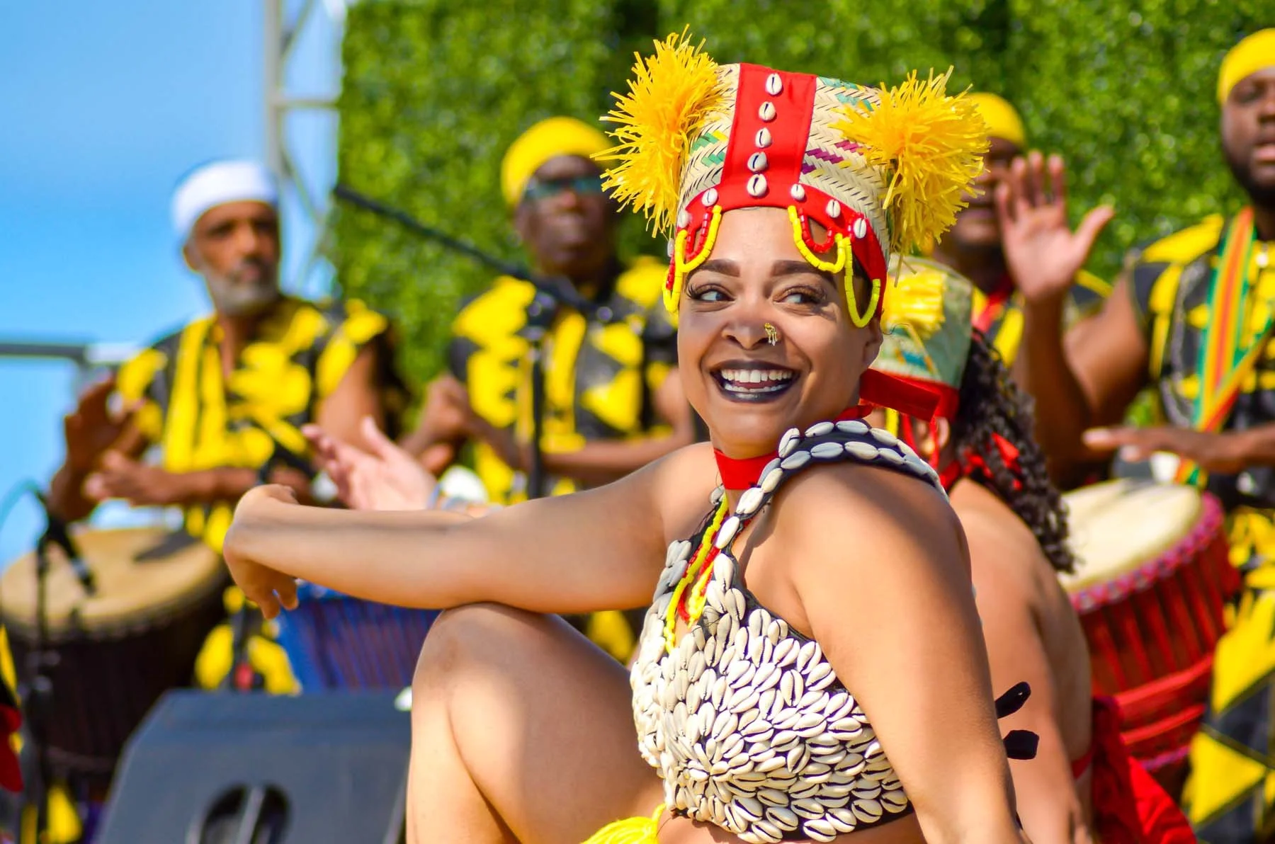 Smiling dancer in a colorful shell costume and feathered headdress performs with drummers at an outdoor cultural festival.