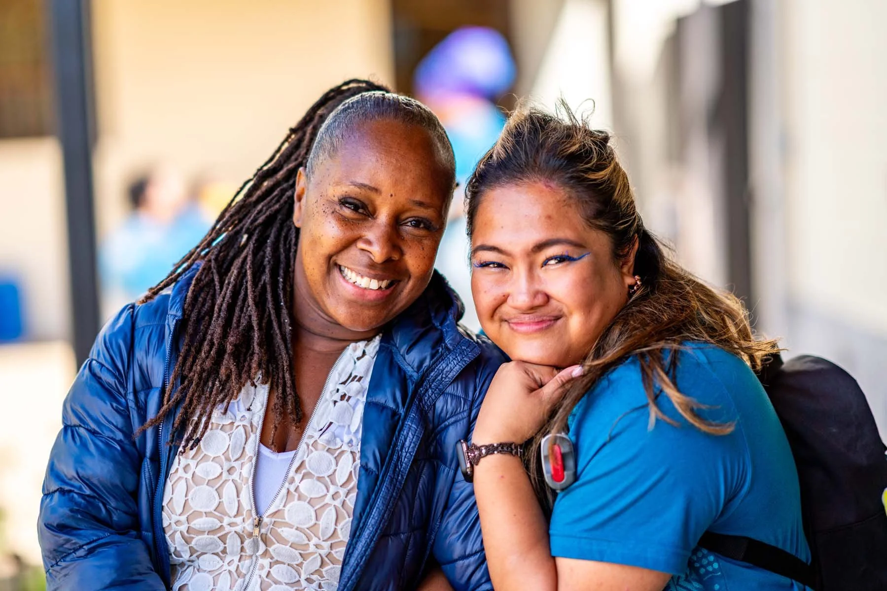 Two smiling women posing together outdoors, one wearing a blue jacket and the other wearing blue scrubs.