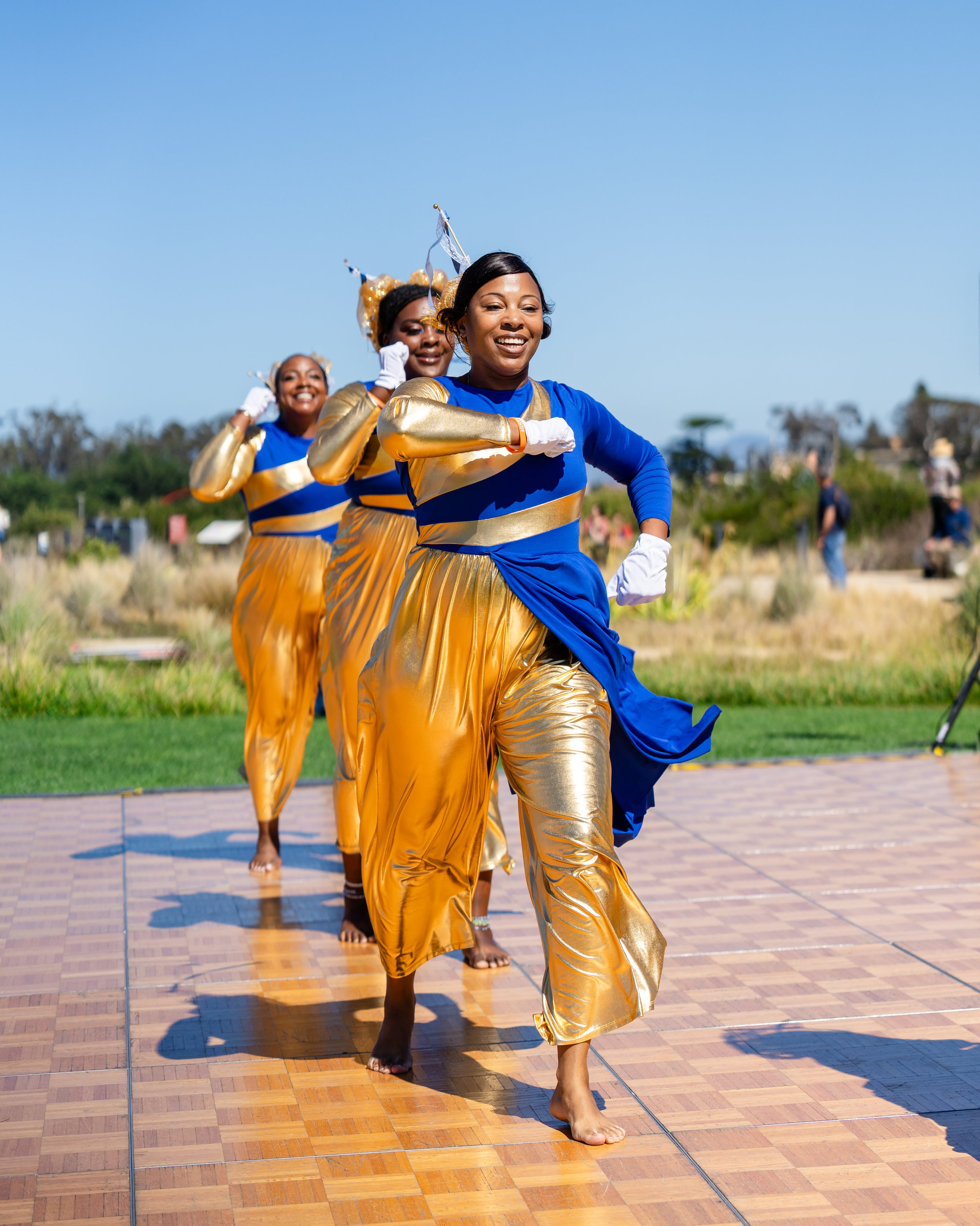 Smiling dancers in blue and gold costumes perform outdoors on a sunny day.