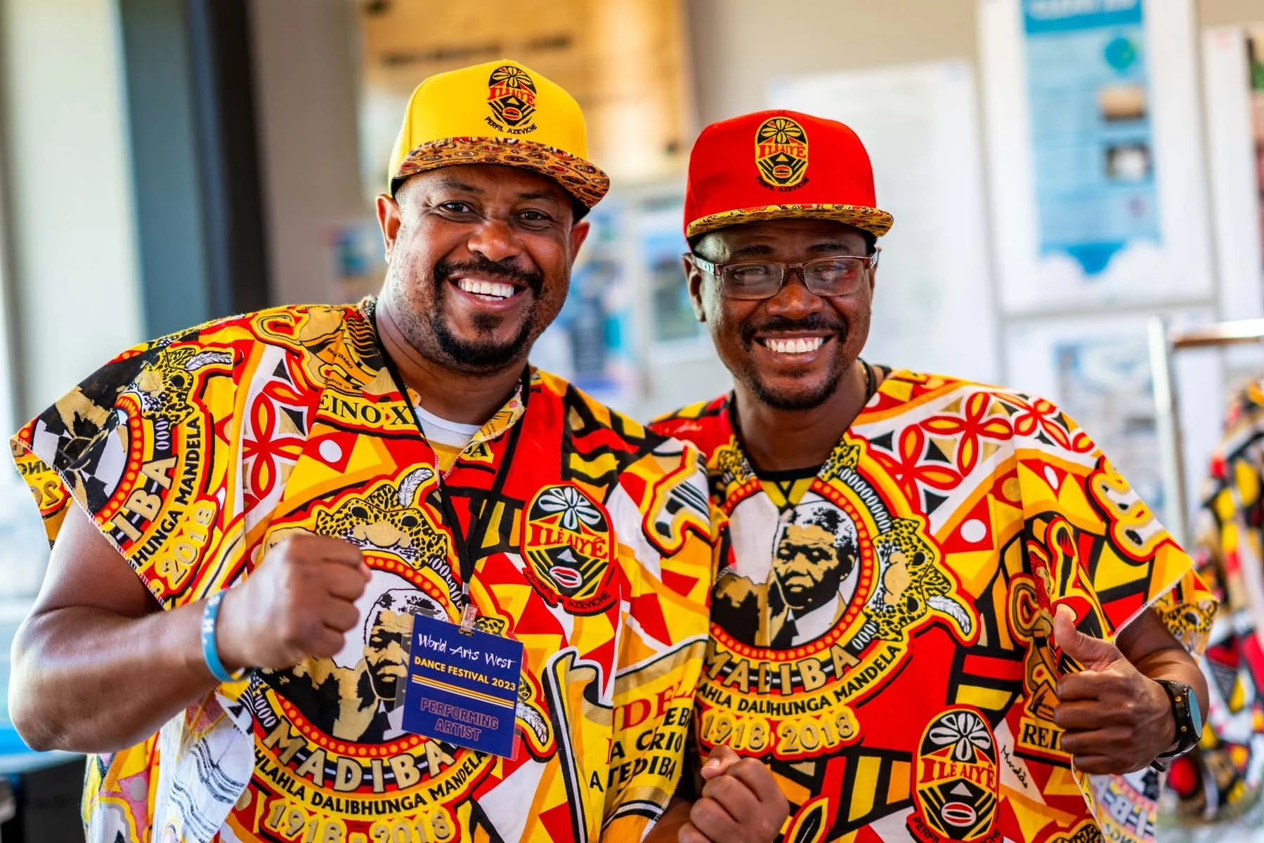 Two smiling men wearing colorful matching African print shirts and caps pose at a dance festival.