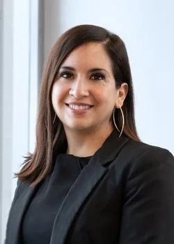 Professional headshot of a smiling woman with long dark hair wearing a black blazer