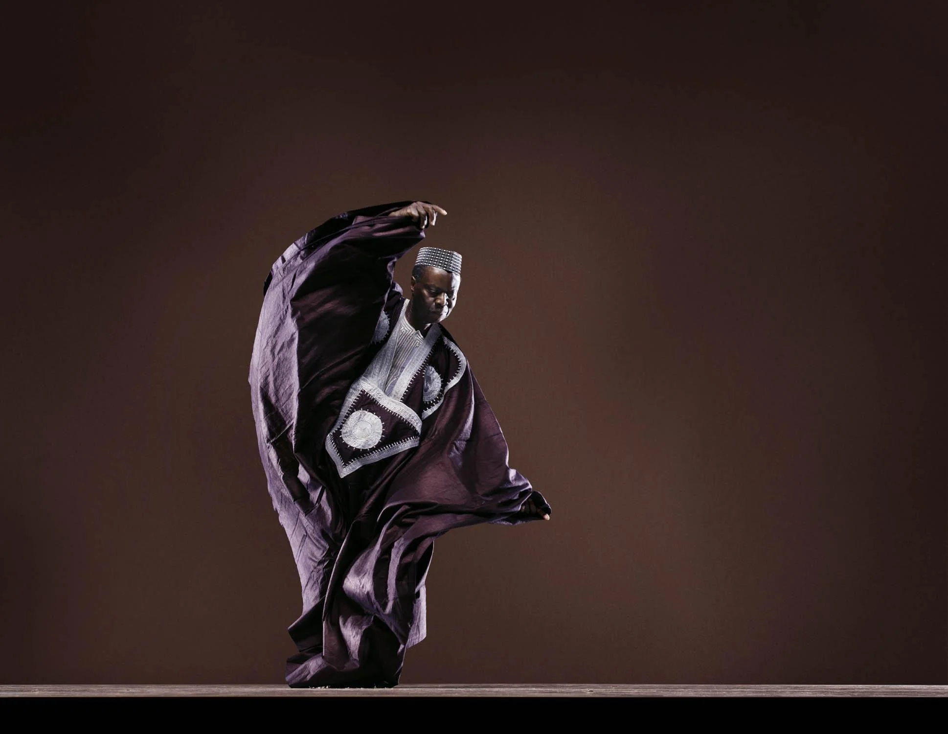 Dancer in embroidered traditional West African clothing performing onstage against a dark brown backdrop
