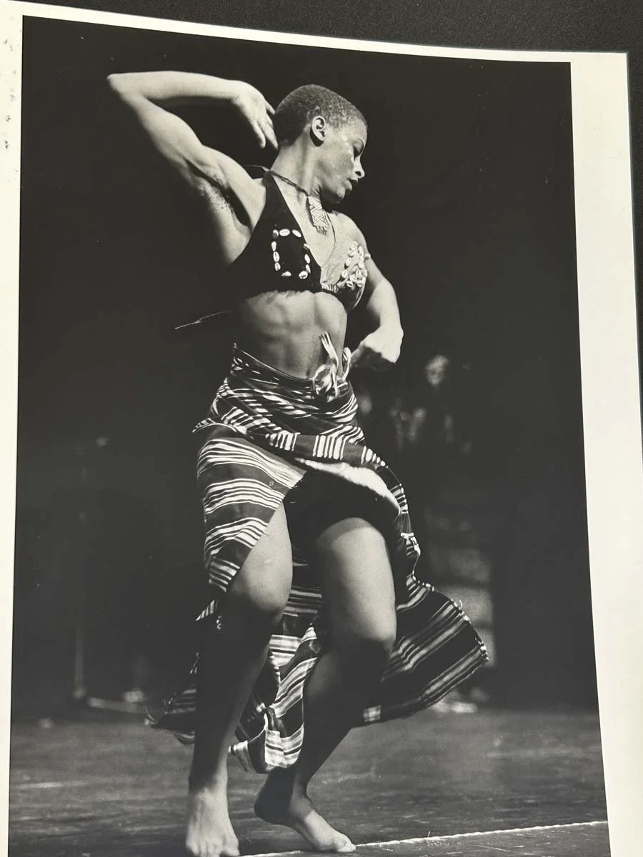 Black-and-white photograph of a dancer performing in a decorated halter top and striped wrap skirt.