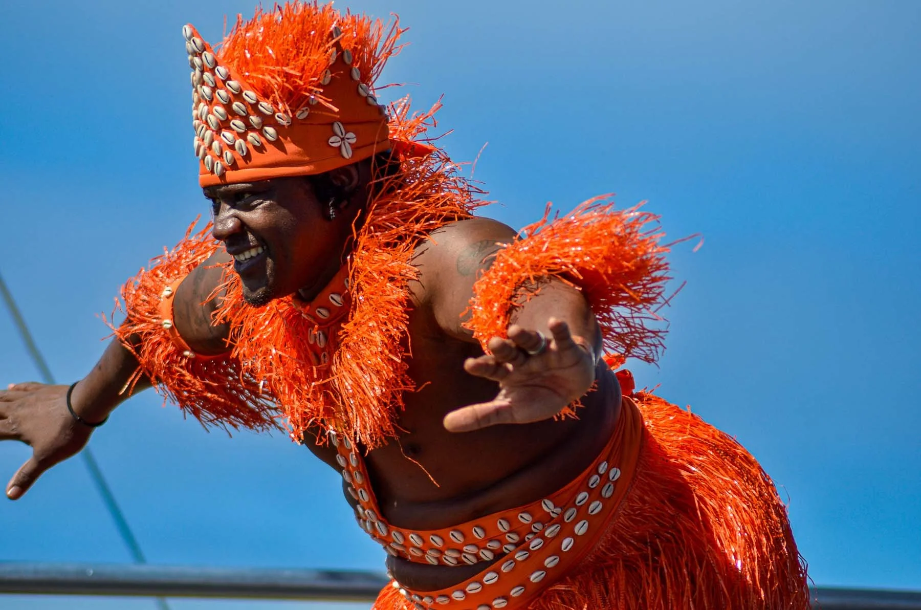 Man in elaborate orange shell-adorned costume and headdress performing against a blue sky