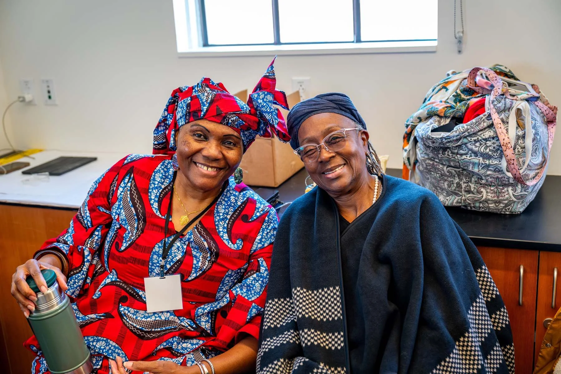 Two smiling older women in colorful African-inspired clothing and headwraps pose indoors, one holding a thermos.