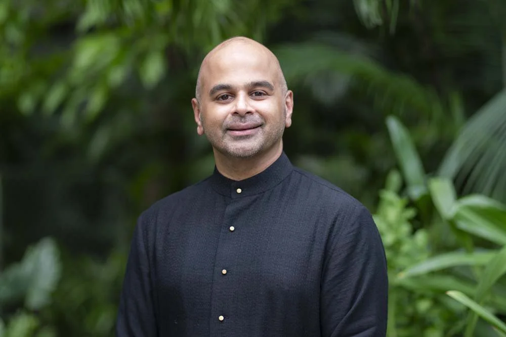 Smiling bald man wearing a black button-up shirt, standing in a lush tropical garden