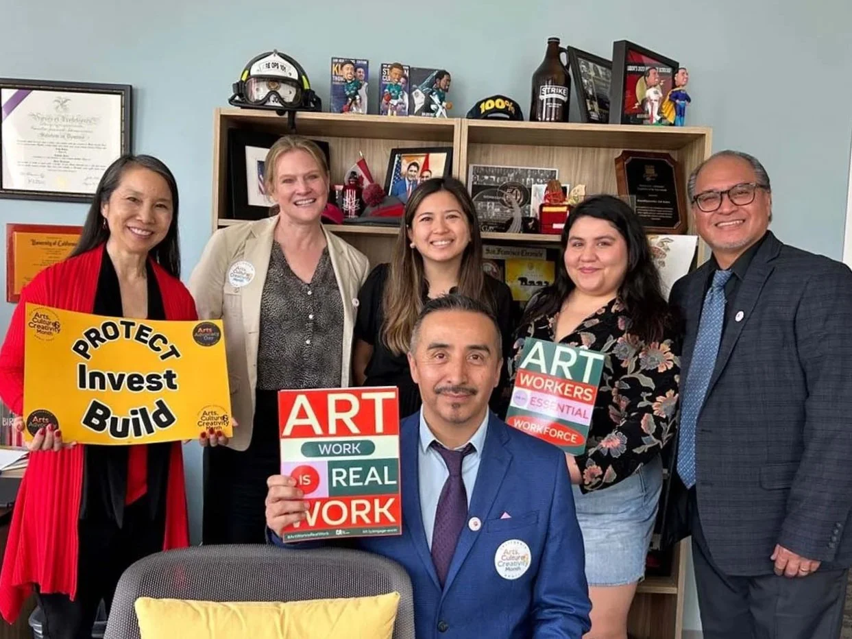 Group of six people holding “Protect Invest Build,” “Art Work Is Real Work,” and “Art Workers Essential Workforce” signs in an arts office