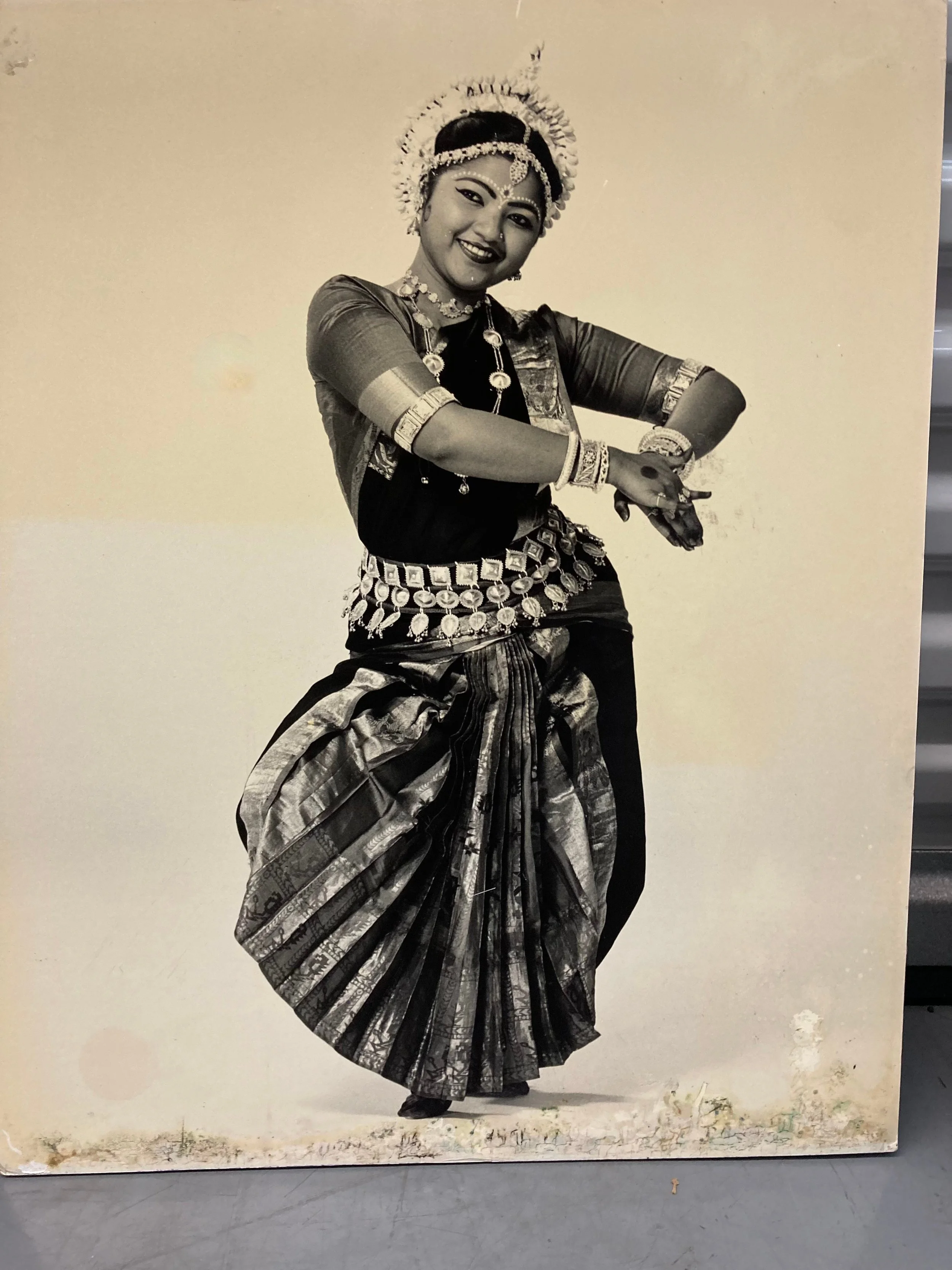 Black-and-white portrait of an Indian classical dancer in an ornate costume, jewelry, and floral headpiece performing a dance pose.