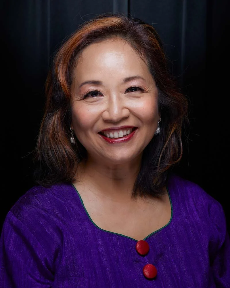 Smiling woman with shoulder-length brown hair wearing a purple blouse against a black background