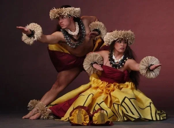 Two Polynesian dancers in traditional grass headpieces, wrist garlands, leis, and colorful costumes performing against a dark studio backdrop.