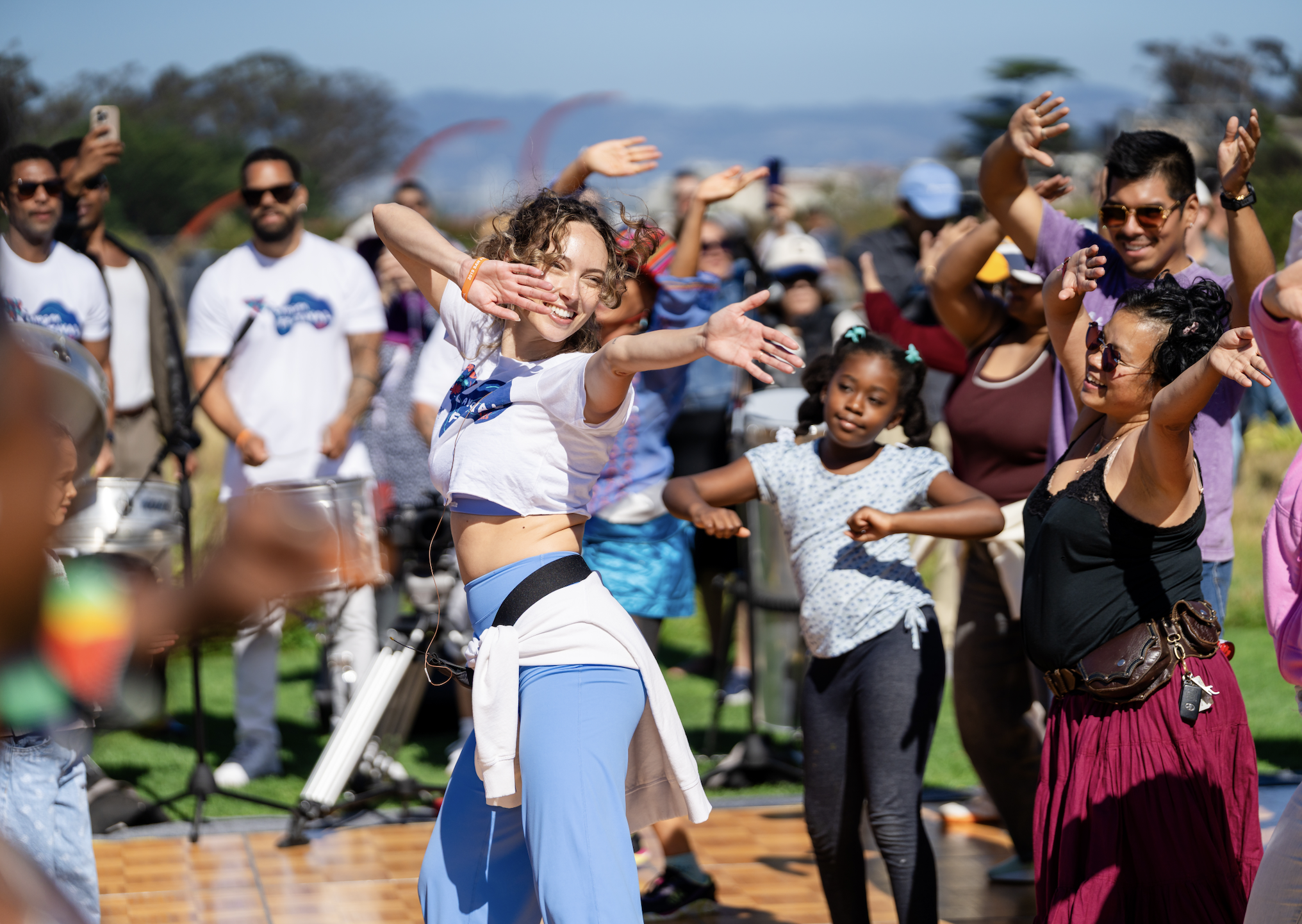 Diverse crowd dancing outdoors at a sunny community event, led by a smiling woman in blue athletic wear.