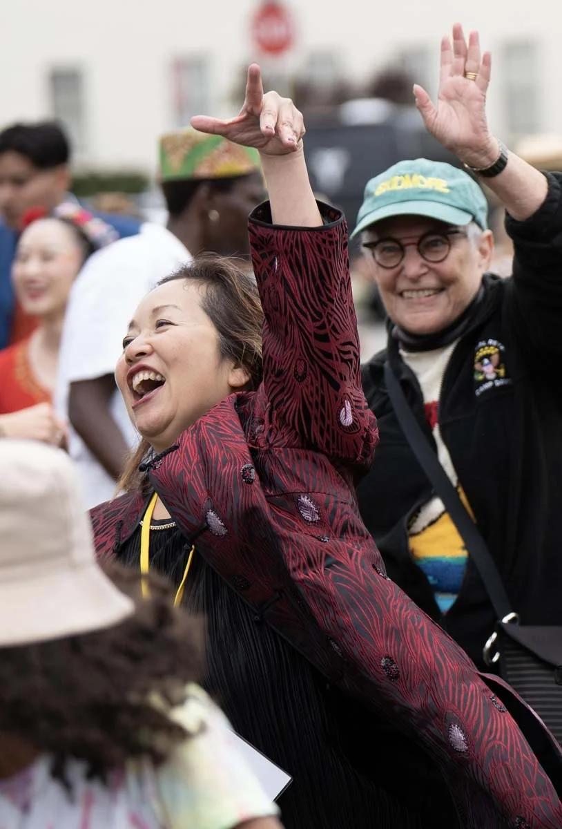 Smiling woman in a patterned red jacket raising her arms in a lively outdoor crowd