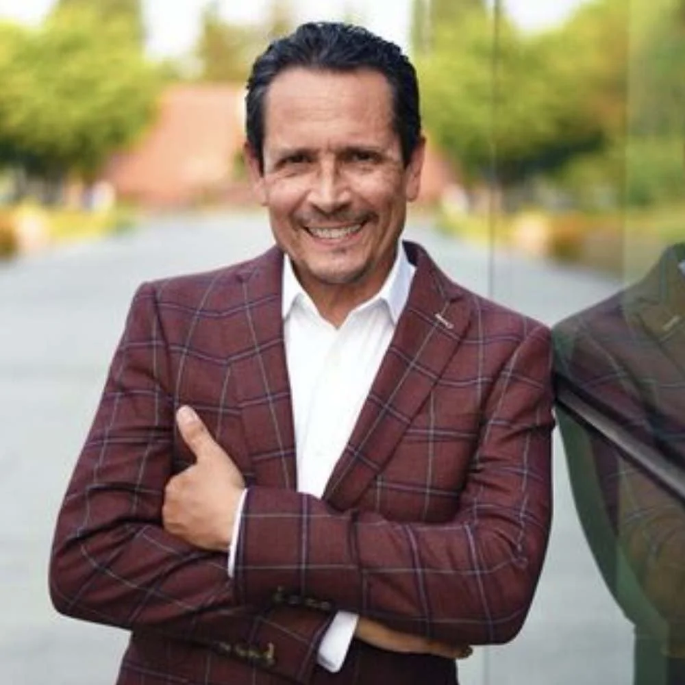 Smiling man in a burgundy plaid suit jacket and white shirt standing outdoors with arms crossed