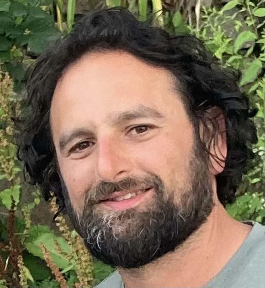 Portrait of a smiling bearded man with dark hair outdoors among green foliage
