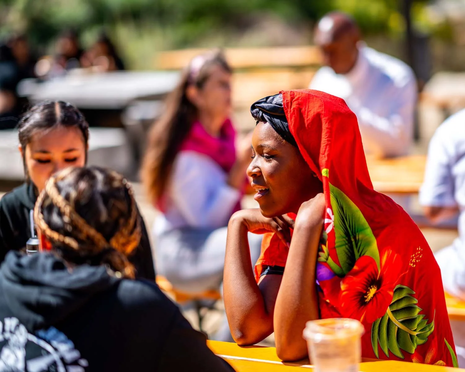 Woman wearing a colorful red floral headscarf sits at an outdoor gathering with other people.