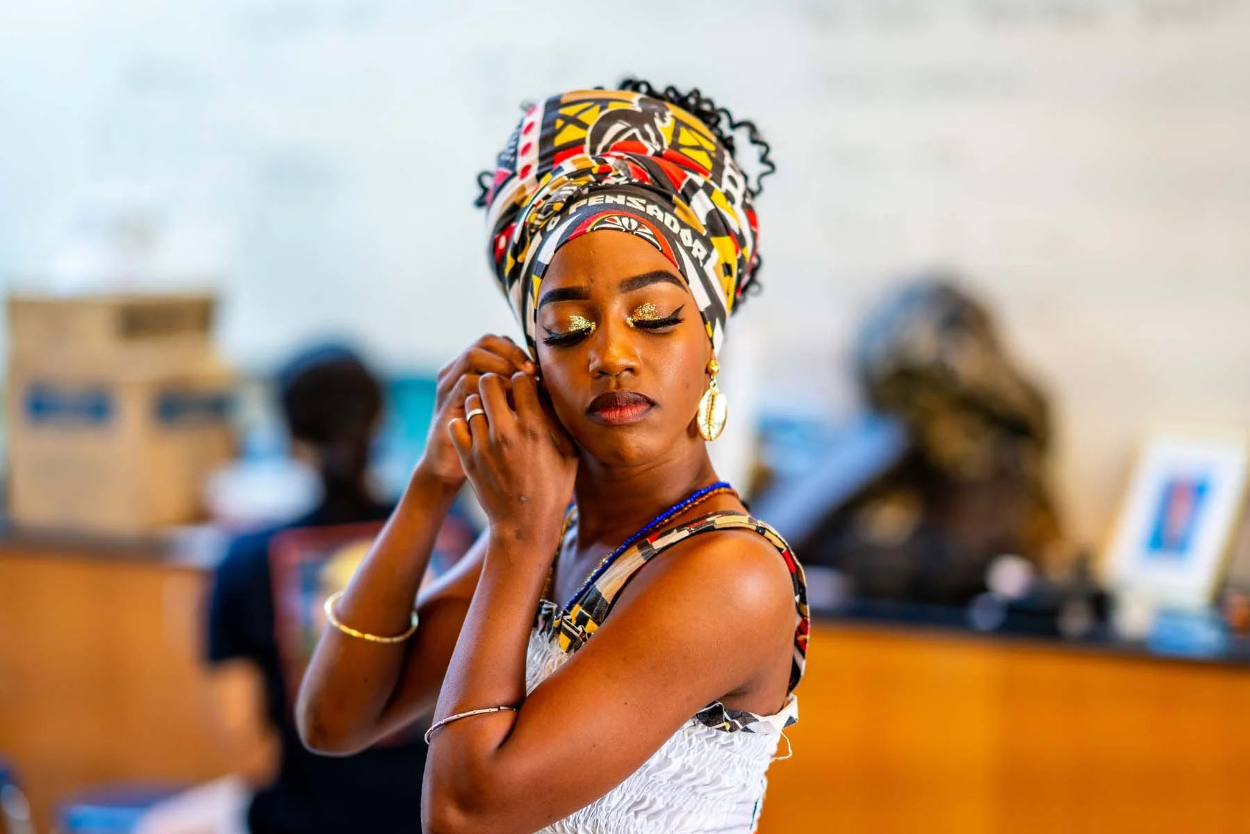 Woman wearing a colorful African headwrap and white dress, adjusting an earring in an indoor setting