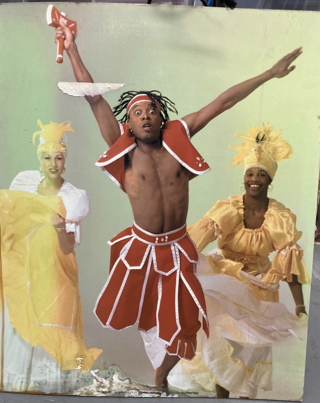 Three costumed dancers perform, with a male dancer in red and white center stage and two women in yellow dresses and feathered headdresses behind him.