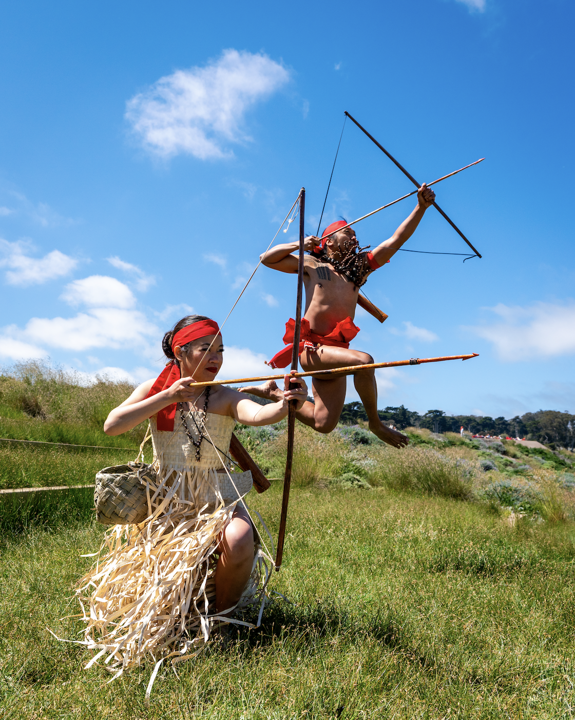 Two performers in traditional Polynesian-inspired clothing demonstrate archery with bows and arrows in a sunny grassy field.
