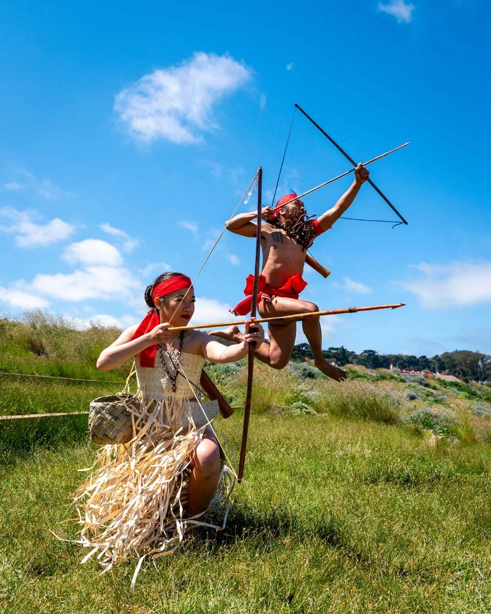 Indigenous performers in traditional clothing reenact archery outdoors beneath a bright blue sky.