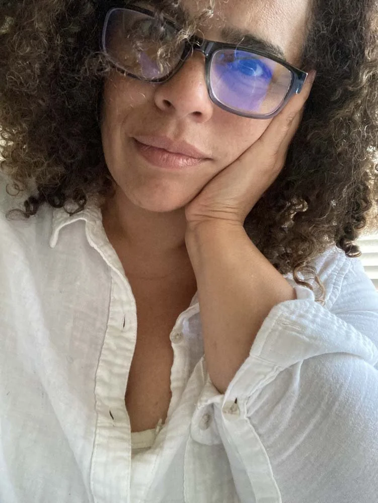 Close-up portrait of a woman with curly hair and glasses wearing a white shirt, resting her face on her hand.