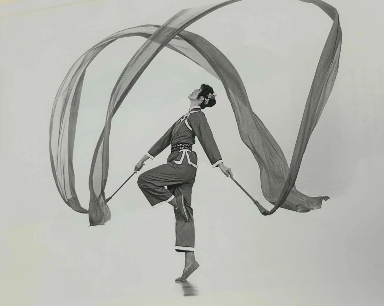 Black-and-white image of a traditional dancer balancing on one foot while waving long silk ribbons.