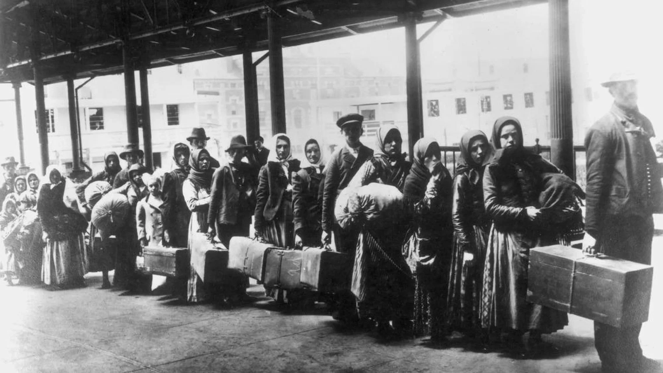 Black-and-white historical photograph of immigrants waiting with luggage at a covered train station platform.