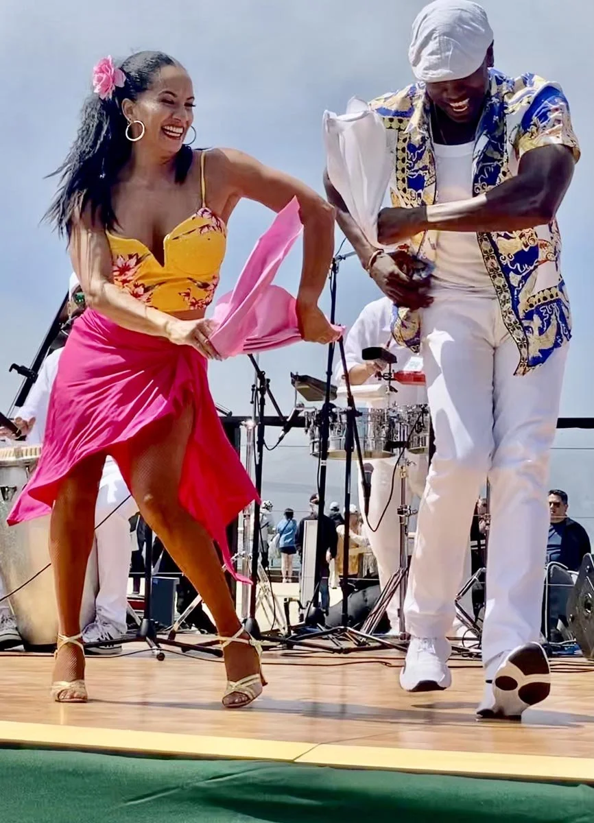 Two performers dancing on an outdoor stage, with a smiling woman in a yellow floral top and pink skirt beside a man in white clothing and a patterned shirt.