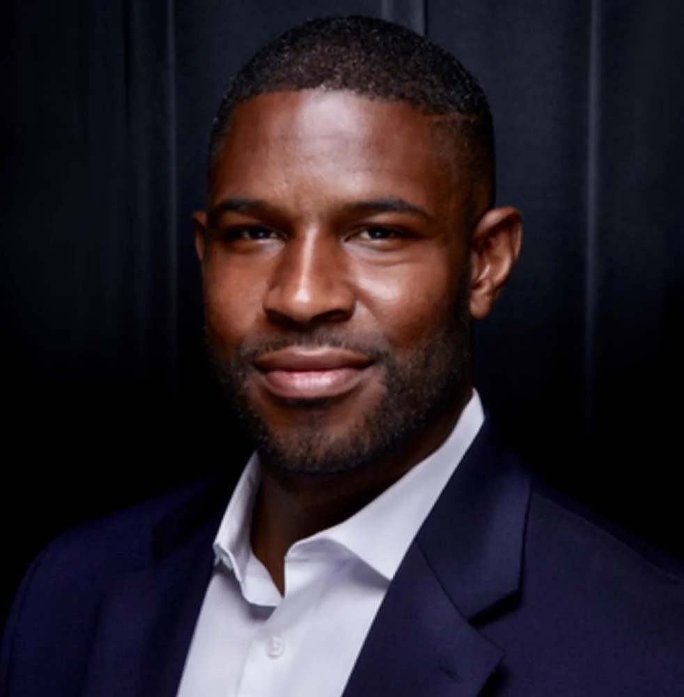 Portrait of a smiling Black man in a navy suit and white dress shirt against a dark background